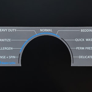 May include: A gray appliance control panel with a curved blue indicator and text labels for wash cycles like "Heavy Duty," "Normal," and "Delicates." The panel also features "Steam Cycles."