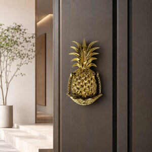 May include: A polished gold-toned metal door knocker shaped like a pineapple. The pineapple features a textured, diamond-patterned body with a leafy crown at the top. A hinged, curved metal striker bar is attached to the sides of the pineapple.