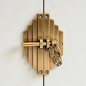 May include: A brass-toned metal door latch featuring a stepped, vertical bar design. The hardware includes a horizontal sliding bolt mechanism and a short, attached security chain that connects to a small post on the right side of the mounting plate.