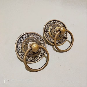 May include: Two ornate brass door pulls with intricate floral designs and ring handles. The pulls are a warm gold color and have a vintage aesthetic.