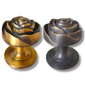 Op de afbeelding: Twee decoratieve kastknoppen met een rozenontwerp. De knop links is messingkleurig en de knop rechts is zwart.