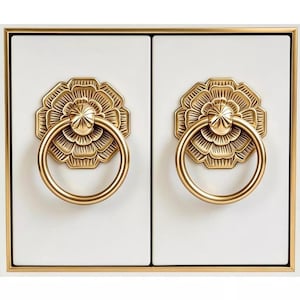 May include: Two white rectangular panels framed in gold, each featuring a centred gold door knocker with a circular ring attached to a decorative floral-shaped backplate. The design is symmetrical, with clean lines and a polished metallic finish.