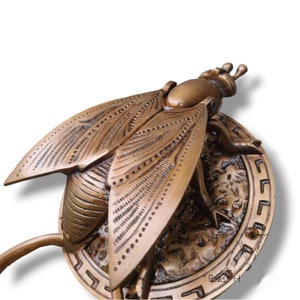 May include: A brass door knocker in the shape of a cicada with intricate details. The cicada is perched on a round base with a Greek key design.
