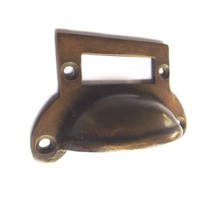 May include: Antique brass drawer pull with a rectangular label holder. The pull has a rounded, slightly curved shape.