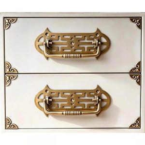 Op de afbeelding: Een ladekast met twee lades in een gebroken witte kleur, voorzien van decoratief beslag in messingkleur. Elke lade is uitgerust met een rechthoekige handgreep met ingewikkelde uitsparingen en bijpassende hoekstukken op de randen.