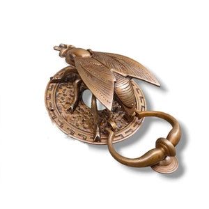 May include: A brass door knocker shaped like a bee with intricate details. The bee is perched on a round base with a decorative border. The knocker has a ring handle.