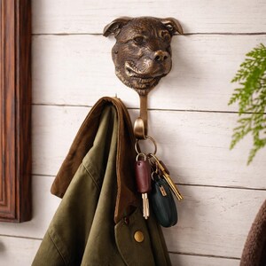 May include: Bronze-colored dog head wall hook with a coat and keys hanging from it. The hook is mounted on a white wooden wall. The coat is olive green with a brown collar. The keys have brown and green keychains.