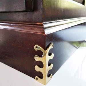 May include: A close-up of a dark brown wooden furniture corner with a decorative brass corner bracket. The bracket has a stylized, organic design. The wood has a polished finish, and the brass has a warm, golden hue.