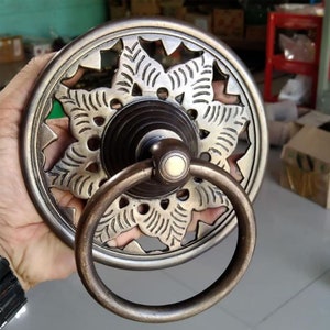 Puede incluir: Un llamador de puerta circular de latón con un diseño floral decorativo y un asa de anillo. El llamador tiene un acabado marrón oscuro y un centro elevado.