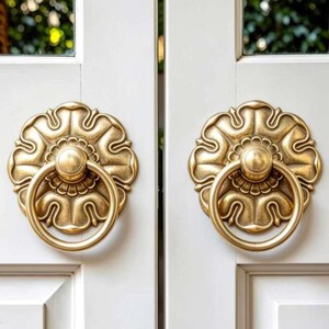 May include: Two matching brass door knockers with a circular ring and a decorative floral-shaped backplate. The hardware features a brushed metallic finish and is mounted on a white panelled door with visible vertical lines and square frames.