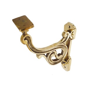 Puede incluir: Un soporte de estante decorativo de metal dorado con un diseño de desplazamiento. El soporte tiene una superficie superior plana y un diseño de desplazamiento decorativo en el lateral.