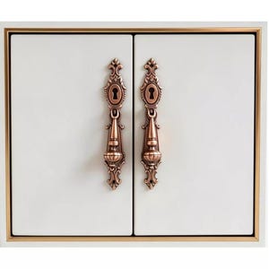 May include: A pair of white cabinet doors framed in gold, featuring two vertical copper-toned metal handles with ornate, scroll-like detailing and integrated keyholes. The handles are centered on each door, providing a contrast to the smooth white surface.