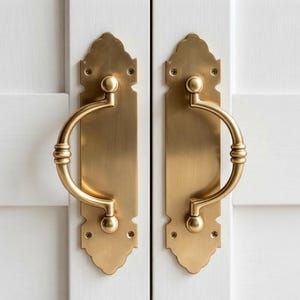 Puede incluir: Dos manijas de puerta de latón a juego con agarres curvos y placas decorativas. Cada manija presenta un acabado en oro cepillado, un tirador semicircular y una placa de montaje de borde festoneado asegurada con cuatro tornillos visibles.