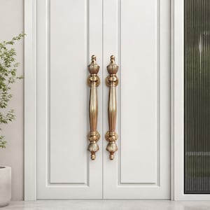 May include: Two ornate, gold-colored door handles are mounted on a white door. The handles have a vertical, elongated design with decorative detailing at the top, middle, and bottom. The door is part of a double door set.