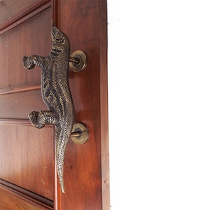Op de afbeelding: Een bronzen deurknop in de vorm van een hagedis met een getextureerd oppervlak. De hagedis klimt de deurknop op en heeft zijn staart om de knop gewikkeld.