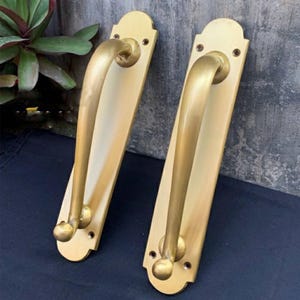 May include: Two antique brass door handles with a curved design and a rounded knob at the end. The handles are mounted on rectangular plates with decorative edges.