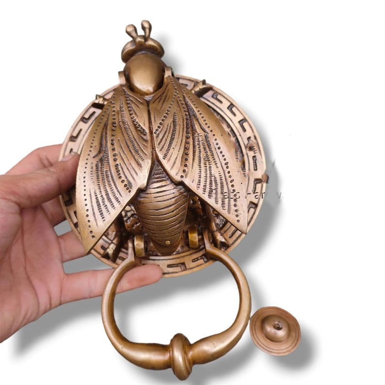 May include: A gold-toned brass door knocker shaped like a stylized fly with intricate details. The knocker has a circular base with a decorative border and a ring handle.