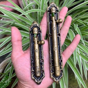 May include: Two antique brass-coloured door bolts with ornate floral designs. The bolts have a rectangular shape with a sliding mechanism and a decorative handle. The bolts are held in a hand against a background of green foliage.