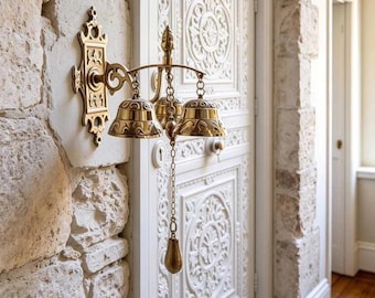 May include: A brass door bell mounted on a stone wall, featuring an ornate rectangular backplate, a curved support arm, and three polished brass bells suspended by chains. A central chain hangs downward with a teardrop-shaped brass pull handle.