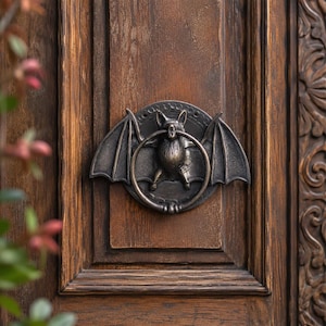 May include: Bronze bat-shaped door knocker with a ring in its mouth, mounted on a dark brown wooden door. The bat has outstretched wings and detailed features. The door has a rectangular panel design.