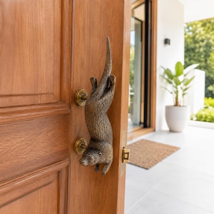 May include: A bronze-coloured metal door handle shaped like an otter, mounted vertically on a wooden door. The otter figure is depicted in a climbing position with its head facing downward and its tail extending upward, secured by two circular mounts.