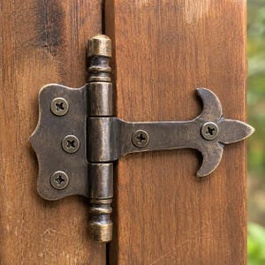 May include: A dark bronze-toned metal T-hinge with a decorative, pointed end and a central cylindrical barrel. The hinge is secured to a vertical wooden surface with five matching screws, each featuring a cross-head drive pattern.