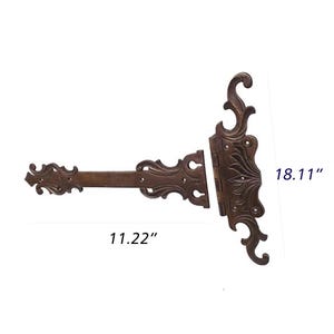 May include: Ornate bronze door hinge with decorative scrollwork and a rectangular bar. The hinge measures 28.5 cm long and 46 cm high. The design features symmetrical patterns and a vintage aesthetic.