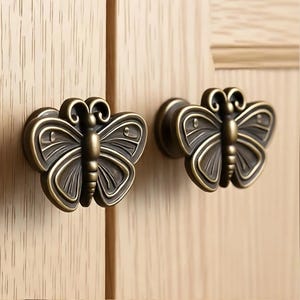 May include: Two butterfly-shaped cabinet knobs made of a dark bronze-toned metal. Each knob features a symmetrical wing design with raised edges and a central body, mounted onto a light-colored wood cabinet door with a visible vertical grain pattern.