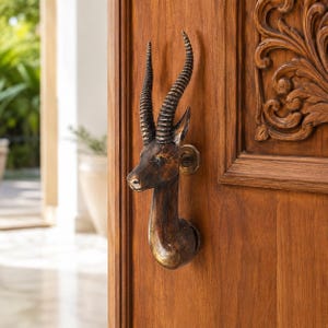 May include: A dark brown, metallic door handle shaped like the head of an antelope with long, spiraled horns. The handle is mounted onto a wooden door featuring carved, ornate floral patterns. The finish on the antelope head is textured.