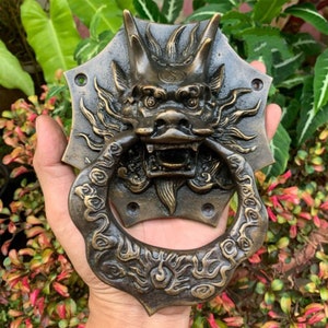 Könnte beinhalten: Ein Türklopfer aus Messing mit einem detaillierten Drachen-Design. Der Drachen hat seinen Mund geöffnet, der scharfe Zähne zeigt, und seine Augen sind weit und wachsam. Der Türklopfer ist kreisförmig, mit einem Ring in der Mitte zum Ziehen.