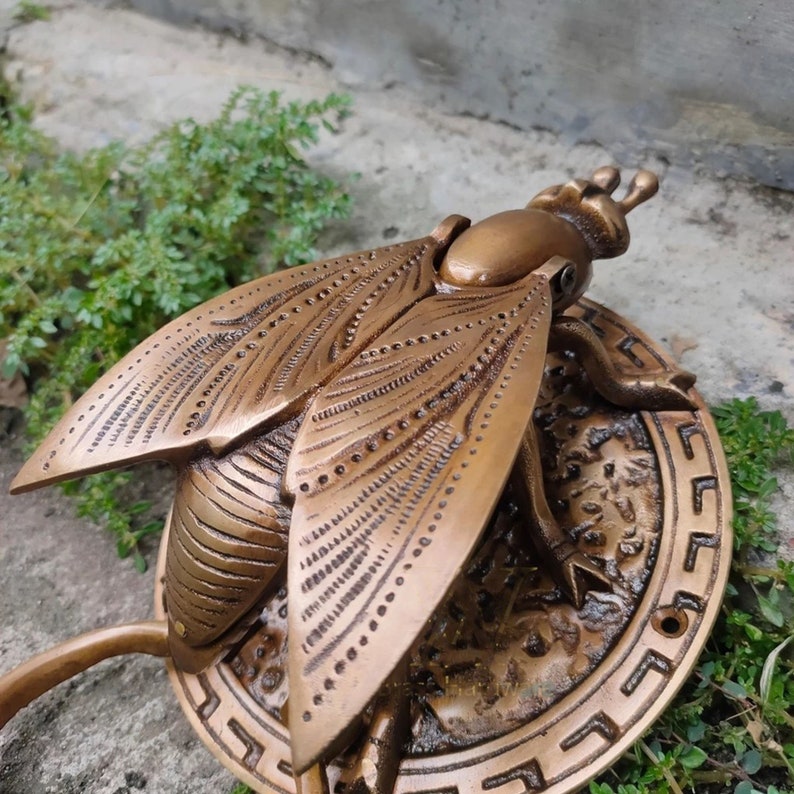 May include: A brass door knocker in the shape of a stylized fly with intricate details. The fly is mounted on a round brass plate with a decorative border.