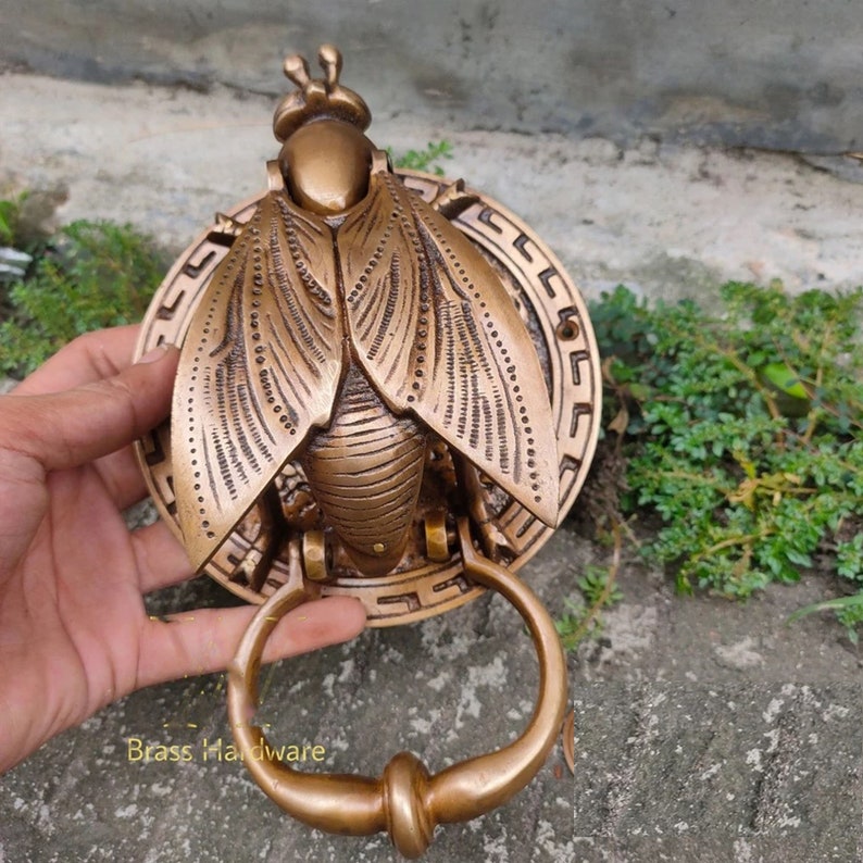May include: A brass door knocker shaped like a fly with intricate details. The fly is mounted on a round plate with a geometric design. The knocker has a ring handle.
