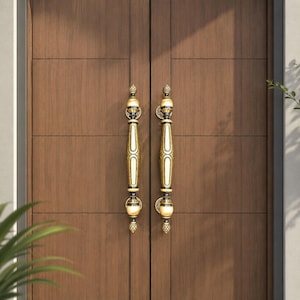 May include: A close-up of a wooden door with two ornate, gold-colored door handles. The handles are vertically oriented, with a detailed design and a decorative finial at the top and bottom. The door is a warm brown color.