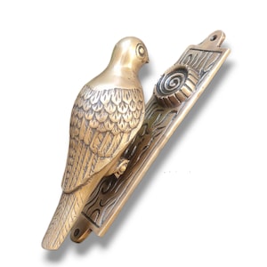 May include: A brass door knocker shaped like a bird with detailed feather markings. The bird is perched on a rectangular plate with a decorative swirl design.