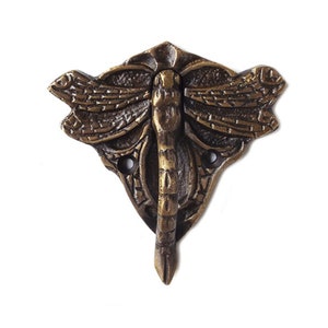 Puede incluir: Un gancho de libélula de metal dorado con detalles intrincados. El gancho tiene forma de libélula con las alas extendidas. Tiene un solo gancho en la parte inferior para colgar objetos.