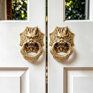 Könnte beinhalten: Zwei passende Türklopfer aus Messing an einer weißen Doppeltür. Jeder Klopfer zeigt ein detailliertes Drachenkopf-Design mit einem kreisförmigen Ring, das durch aufwendige Reliefmuster und eine polierte Metalloberfläche der Beschläge besticht.