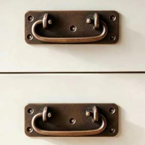 May include: Two rectangular bronze-toned metal drawer pulls with a dark, weathered finish. Each handle features a central swinging bail attached to a flat, four-hole mounting plate. The hardware is mounted horizontally on a smooth, off-white surface.