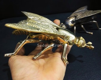 May include: Two brass fly shaped cigarette lighters. The lighter on the left is gold and the lighter on the right is a darker brass color.