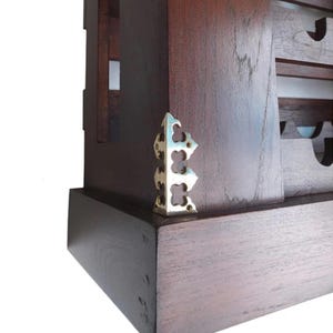 May include: Close-up of a dark brown wooden cabinet corner with a decorative brass corner bracket. The bracket has a stylized, openwork design. The cabinet appears to be part of a larger piece of furniture.