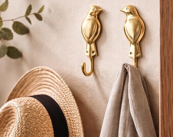 May include: Two gold-colored bird-shaped wall hooks. The hooks are mounted on a light-colored wall, with a straw hat and a light gray scarf hanging below. The hooks are made of metal and have a curved hook shape.