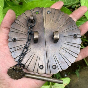 Könnte beinhalten: Ein Türverschluss aus Messing mit einem dekorativen Sonnenstrahlen-Design und einem Schlüsselloch. Der Verschluss hat eine Kette daran und einen Schlüssel.