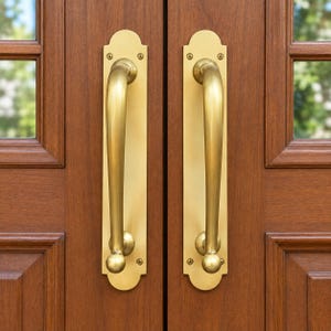 May include: A pair of matching brass door handles mounted on a wooden door. Each handle features a long, curved grip attached to a rectangular backplate with rounded ends, secured by four visible screws at the top and bottom corners.