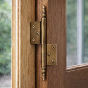 May include: Close-up of a brass door hinge with decorative detailing. The hinge is mounted on a light brown wooden door frame. The hinge has a rectangular plate with three screws on each side, and a cylindrical central section.