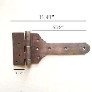 Puede incluir: Una bisagra en T de metal oscuro con acabado desgastado, que presenta múltiples orificios para tornillos. La bisagra tiene una longitud total de 28,98 cm, una longitud de correa de 22,48 cm y un ancho de placa de 4,50 cm según el texto.