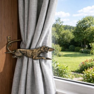 May include: A bronze-colored metal curtain tieback in the shape of a dog, attached to a wooden door frame. The dog is in a reclining position, holding a gray curtain. The background shows a window with a view of a green lawn and trees.