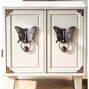 May include: A small, off-white cabinet with two doors, featuring dark bronze butterfly-shaped door pulls with integrated circular ring handles. The cabinet has decorative bronze corner accents on the top and bottom edges of the front frame.