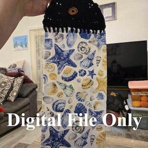 May include: A white tea towel with a blue and yellow seashell pattern. The tea towel has a black crocheted hanging loop with a wooden button.