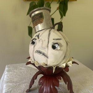 May include: A white ceramic pumpkin with a painted face and a silver metal top hat. The pumpkin is sitting on a red metal stand with three legs.