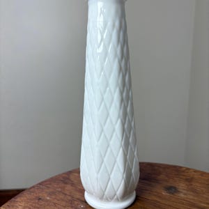 May include: A tall, white, diamond-patterned vase with a scalloped rim. The vase is made of a glossy material and has a flared shape, standing on a small, round base. The vase is approximately 10 inches tall.