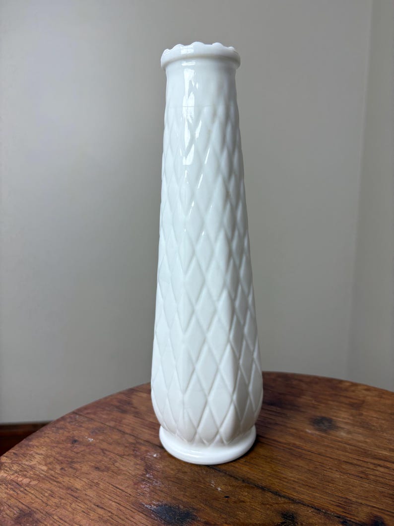 May include: A tall, white, diamond-patterned vase with a scalloped rim. The vase is made of a glossy material and has a flared shape, standing on a small, round base. The vase is approximately 10 inches tall.